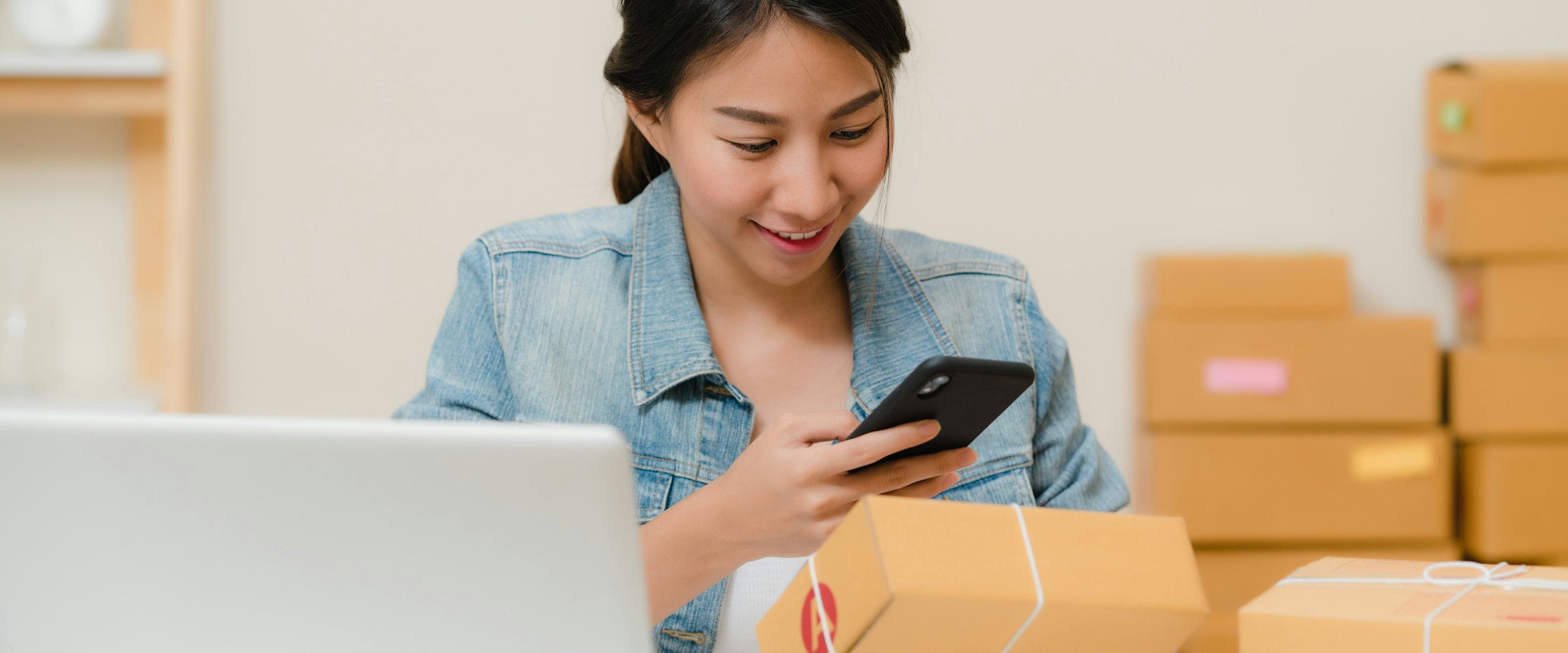 Webshop-eigenaar controleert een bestelling op haar telefoon, met pakketten op de achtergrond